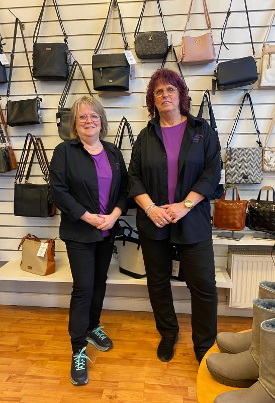 Två kvinnor i svarta kläder och lila skjortor står i en butik framför en skyltvägg med handväskor; stövlar och fler väskor finns på hyllorna nedanför.