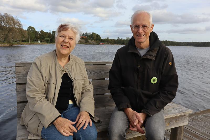 Två äldre vuxna sitter på en träbänk vid en sjö, båda ler mot kameran, med träd och vatten i bakgrunden.