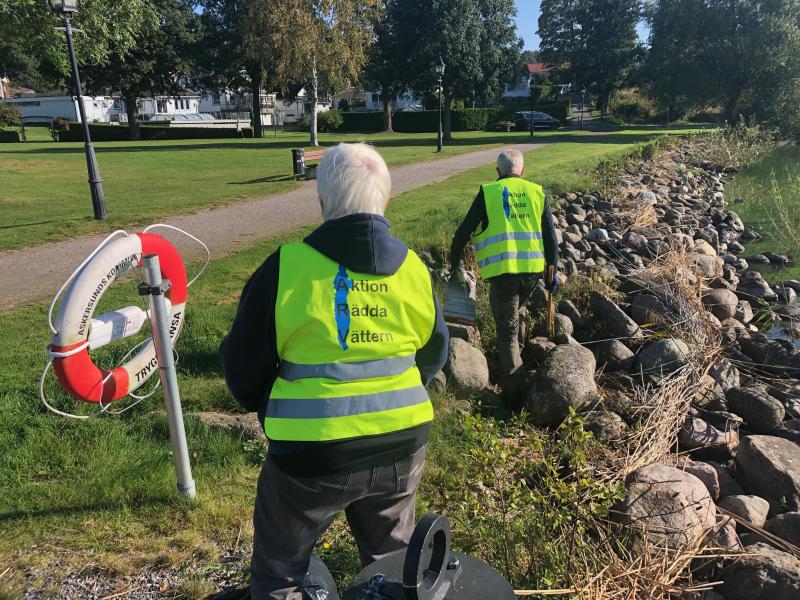 Två personer i gula säkerhetsvästar går längs en klippig strand nära en gräspark, med en livboj synlig till vänster.