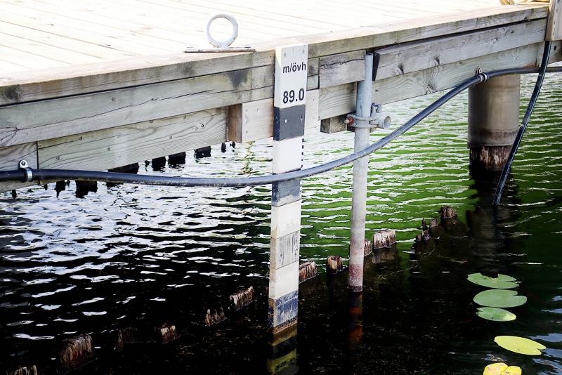 En brygga ligger ovanför vattenytan med en mätstång märkt ”89,0” m, några kablar och näckrosblad som flyter i närheten.
