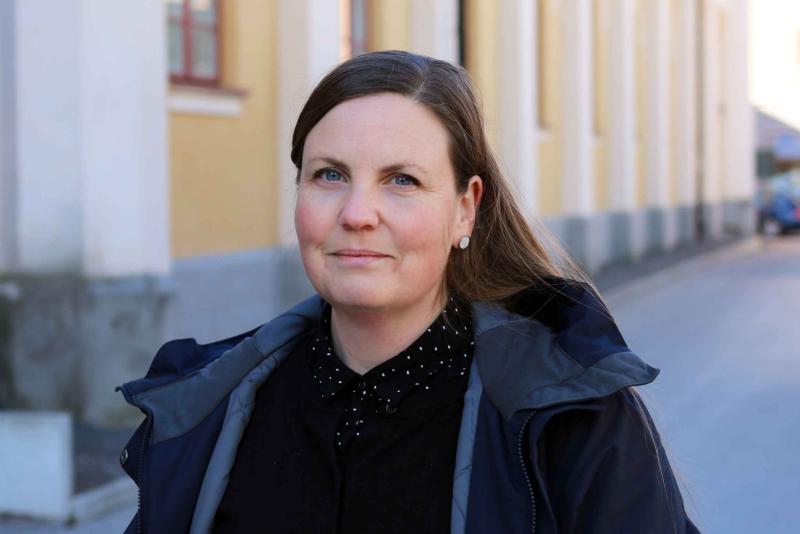 En kvinna med mörkt hår tillbakadraget, iklädd en svart jacka och en prickig skjorta, står utomhus med byggnader i bakgrunden. hon stirrar lite åt höger med ett neutralt uttryck.