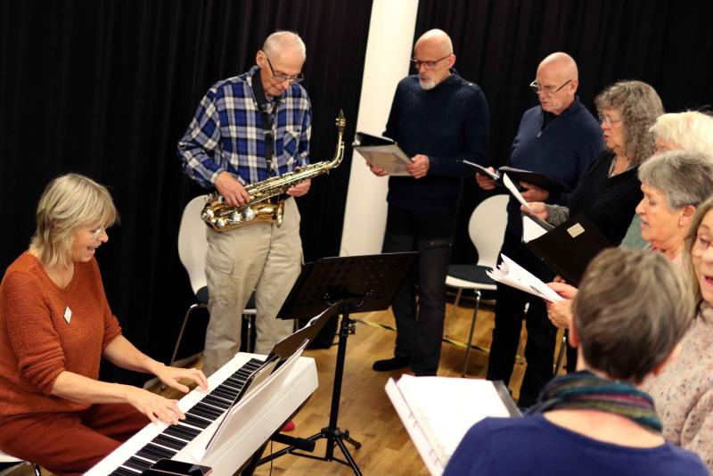 En grupp vuxna musiker och sångare övar tillsammans, med en kvinna som spelar piano i förgrunden och andra håller noter, inklusive en man med saxofon.
