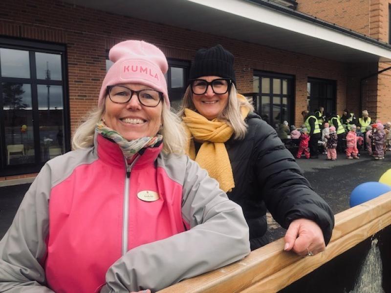 Två kvinnor i vinterkläder ler mot kameran utanför en tegelbyggnad, med en grupp små barn i färgglada kläder i bakgrunden.