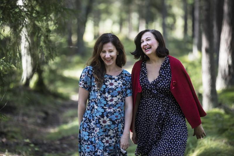 Två kvinnor som skrattar och går tillsammans genom en skog, en i en blommig blå klänning och den andra i en prickig klänning med en röd kofta.