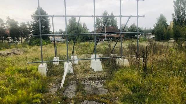 En metallram för en skylt står tom i en övervuxen gräsyta med byggnader och träd i bakgrunden under en molnig himmel.