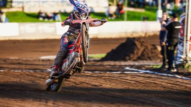 En motocrossförare gör en wheelie på en grusbana under ett lopp, med åskådare och teammedlemmar synliga i bakgrunden.