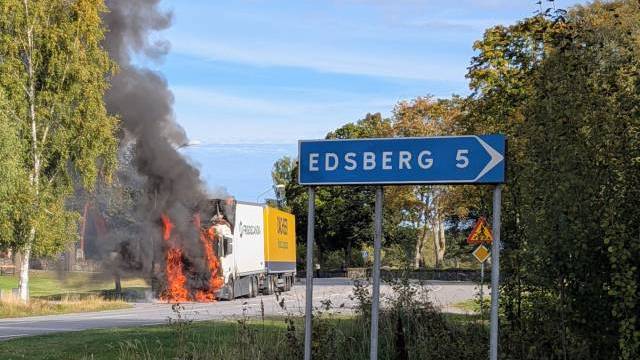 En lastbil som står i brand vid vägkanten sprider svart rök nära en vägskylt som anger att Edsberg ligger 5 kilometer bort.