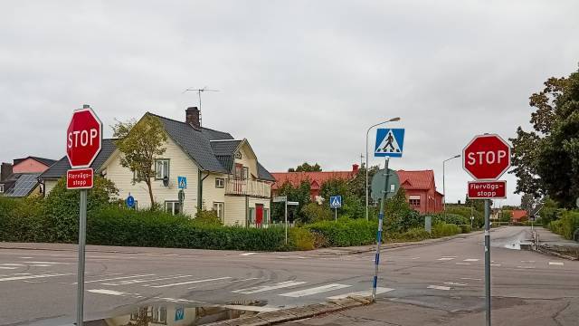 Två stoppskyltar och en övergångsskylt vid en korsning i ett bostadsområde med hus, grönska och molnigt väder.