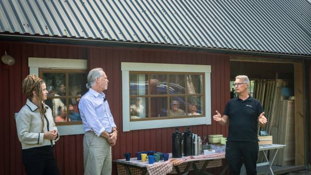 Tre personer står och pratar utanför en röd träbyggnad nära ett bord med muggar, termosar och tallrikar, medan andra syns i fönstret bakom dem.