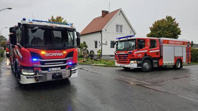 Två brandbilar är parkerade på en våt gata nära ett vitt hus, med flera brandmän som står vid byggnaden under molnig himmel.