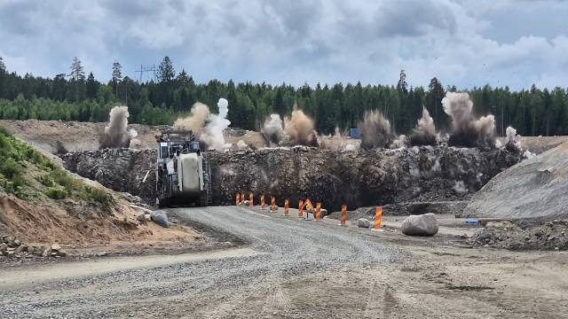 En byggarbetsplats med en grusväg, orange avspärrningar, en stor maskin och en explosion som spränger stenar i bakgrunden, med en skog och ett molnigt himmel.