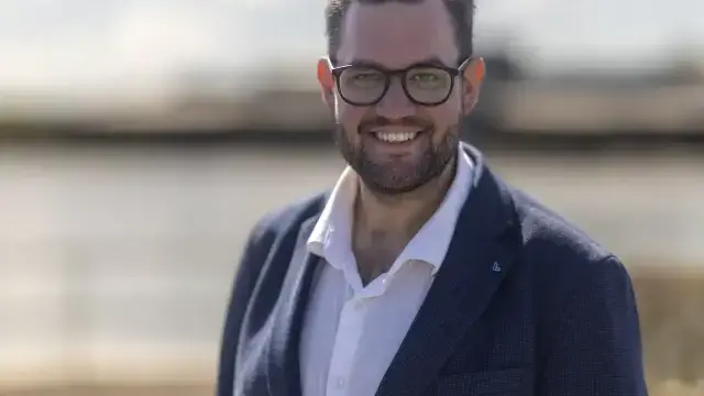 En man med glasögon och blå kavaj över en vit skjorta står utomhus och ler mot kameran med en suddig bakgrund.