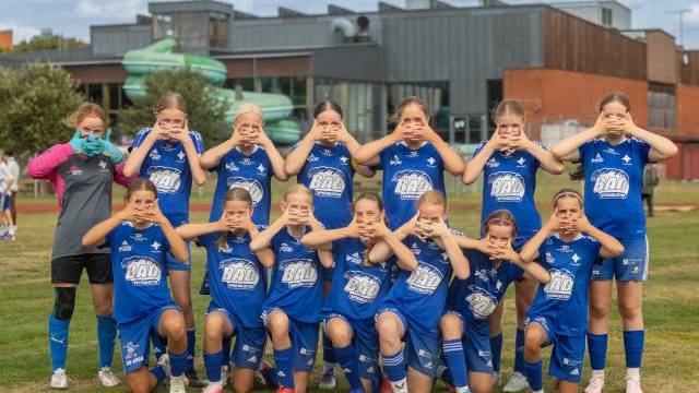 Ett flickfotbollslag i blåa tröjor poserar på en plan, där varje spelare täcker ögonen, öronen eller munnen med händerna. I bakgrunden syns en byggnad och träd.