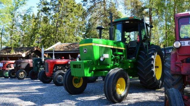 En rad veterantraktorer, däribland en grön John Deere 4250, står parkerade utomhus på en grusplan med träd och träbyggnader i bakgrunden.