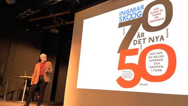 En person står på scenen bredvid ett stort projicerat bokomslag med titeln 