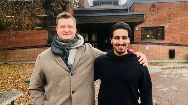 Två män står sida vid sida, den ene i beige kappa och halsduk, den andre i svart huvtröja, och ler framför en tegelbyggnad med höstlöv på marken.