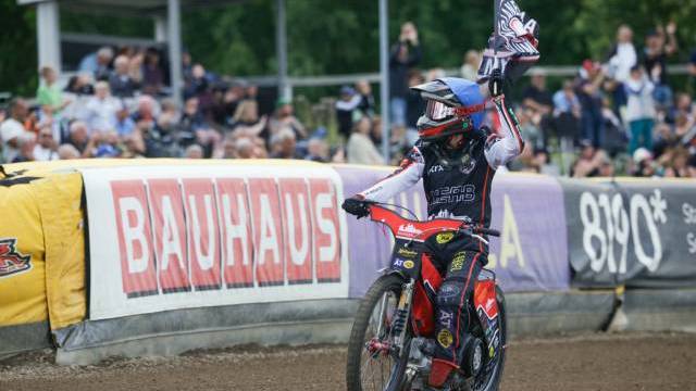 En speedwayförare på en grusbana viftar med en flagga medan han kör förbi publiken, med reklambanderoller synliga i bakgrunden.