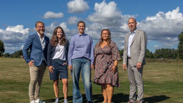 Fem vuxna står utomhus på gräset under en blå himmel med moln, klädda i vardagskläder och affärskläder, och tittar in i kameran och ler.