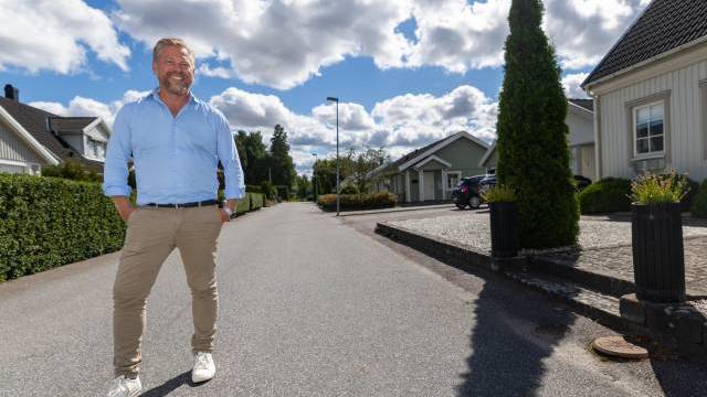 En man i en ljusblå skjorta och beige byxor står och ler på en lugn bostadsgata kantad av hus och grönska under en delvis molnig himmel.