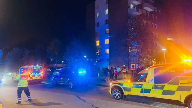 Räddningsfordon med blinkande ljus står parkerade utanför ett flerbostadshus på natten, med människor samlade i närheten och en räddningspersonal som dirigerar trafiken.