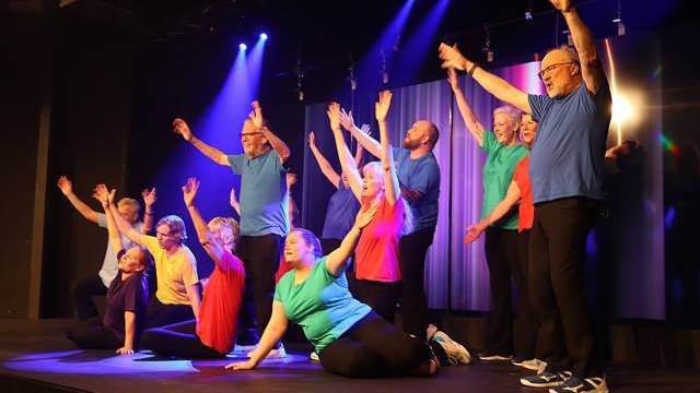 En grupp vuxna på scenen i färgglada skjortor utför en koreograferad rutin med höjda armar under scenbelysning.