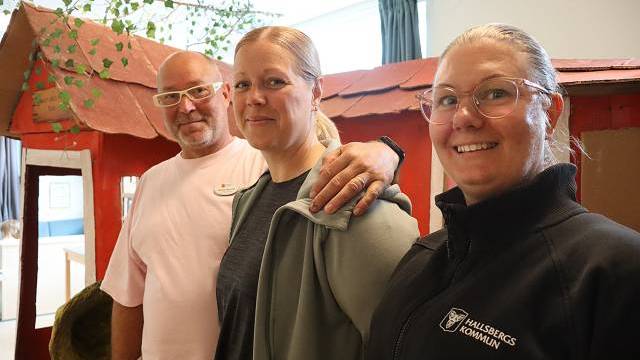Tre vuxna står inomhus framför en liten röd lekstuga; två kvinnor bär glasögon och en man vilar sin hand på den ena kvinnans axel.