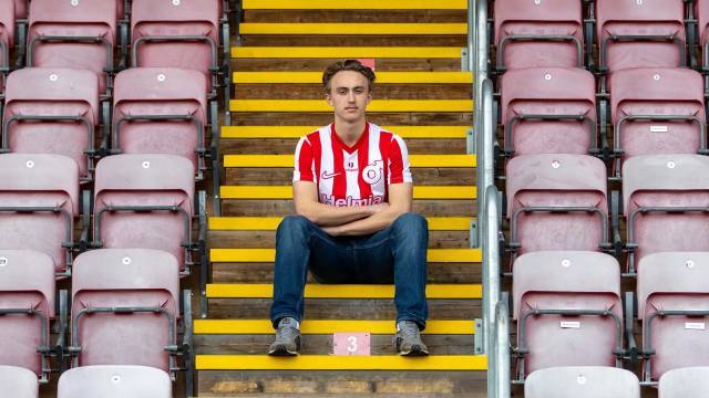 En man i en rödvitrandig fotbollströja sitter ensam på en trappa bland tomma stolar, med armarna i kors och blicken riktad mot kameran.
