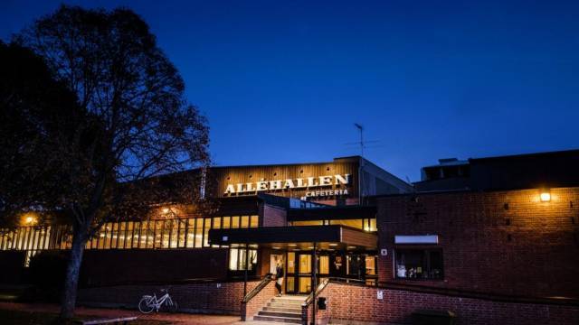 Vy från utsidan av Allehallens cafeteria i skymningen, med tegelväggar, upplyst skylt, träd till vänster och en parkerad cykel nära ingången.
