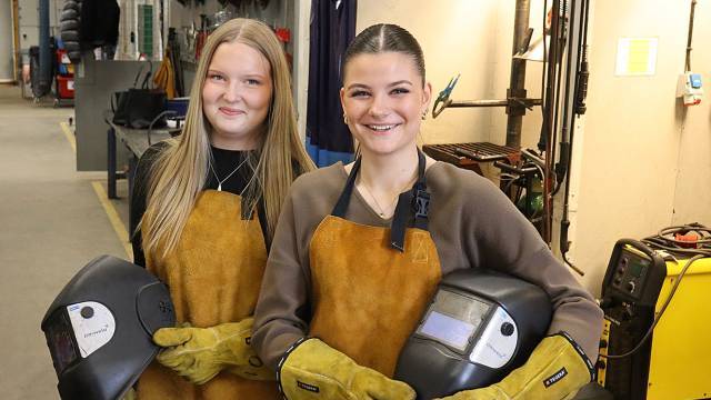 Två kvinnor med svetshandskar och förkläden står inomhus med svetshjälmar och ler mot kameran i en verkstadsmiljö.