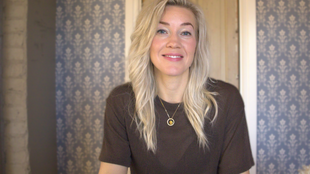 En kvinna med långt blont hår, klädd i en mörk topp och ett hängande halsband, sitter framför en mönstrad blå tapet och en trädörr och ler mot kameran.