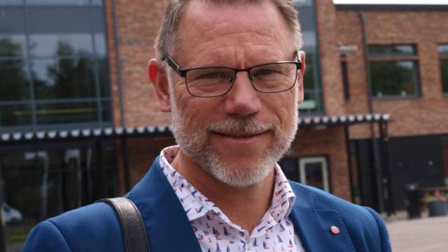 En medelålders man med glasögon och skägg, klädd i blå kavaj och skjorta med tryck, står utanför en modern tegelbyggnad.