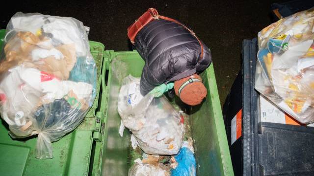 En person klädd i jacka och mössa lutar sig in i en grön soptunna fylld med skräp och rotar igenom påsar mitt i natten. I närheten syns ytterligare soptunnor och soppåsar.