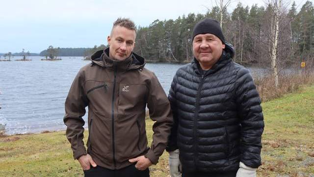 Två kavajklädda män står utomhus vid en sjö med träd och mulen himmel i bakgrunden.