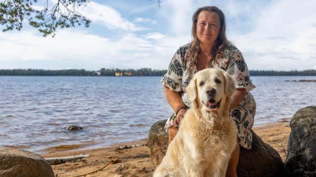 En kvinna i en mönstrad klänning sitter vid en sjöstrand tillsammans med en golden retriever; båda vänder sig mot kameran. I bakgrunden syns träd och vatten under en delvis molnig himmel.