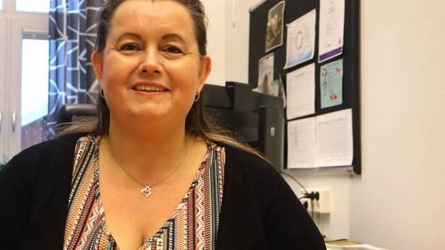 En kvinna med långt brunt hår och en mönstrad topp sitter och ler i en kontorsmiljö med en anslagstavla och papper i bakgrunden.