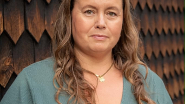 En kvinna med långt, vågigt brunt hår och en grön tröja står framför en vägg med mörka träpaneler.