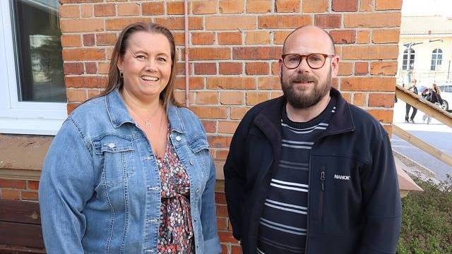 Två vuxna står framför en tegelvägg, den ena i jeansjacka och blommig klänning, den andra i svart jacka och randig skjorta.