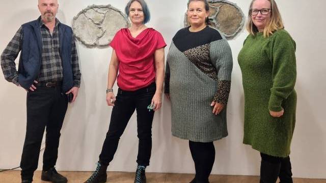 Fyra vuxna står sida vid sida inomhus framför en vit vägg med två abstrakta stenkonstverk. De är vända mot kameran och klädda i avslappnade vinterkläder.