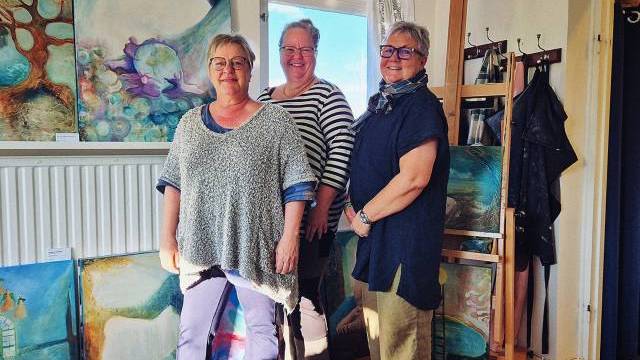 Tre kvinnor står och ler i ett starkt upplyst rum fyllt med färgglada tavlor, bland annat abstrakta verk och naturmotiv, där vissa är utställda på golvet och andra på stafflier och på väggarna.