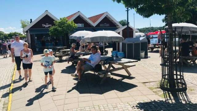 Folk sitter vid picknickbord under parasoller och promenerar längs en solig gågata med små butiker och träd i bakgrunden.