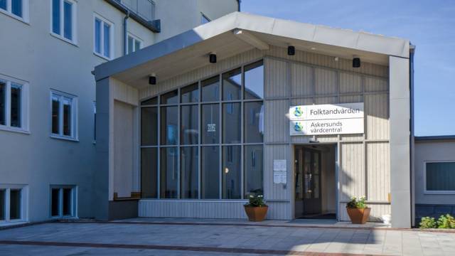 Ingången till en vårdinrättning med stora fönster, krukväxter och skyltar med texterna ”Folktandvården” och ”Askersunds vårdcentral” på ytterväggen.