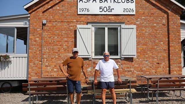 Två män står framför en tegelbyggnad med en skylt där det står ”Aspa Båtklubb 50 år 1976 2026”. Längs väggen står bänkar och på marken ligger grus.
