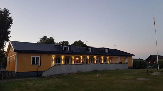 En gul byggnad med mörkt tak, upplyst av ytterbelysning i skymningen, står på en gräsmatta med en flaggstång till höger.