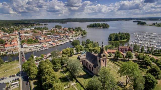 Flygfoto över en stad vid en sjö med en kyrka, marina, båtar och kluster av byggnader omgivna av gröna träd under en delvis molnig himmel.