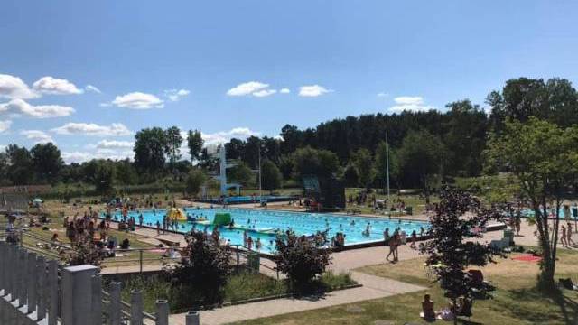 Allmän utomhuspool en solig dag med många människor som simmar, slappar och promenerar runt poolområdet; träd och gräs omger anläggningen.