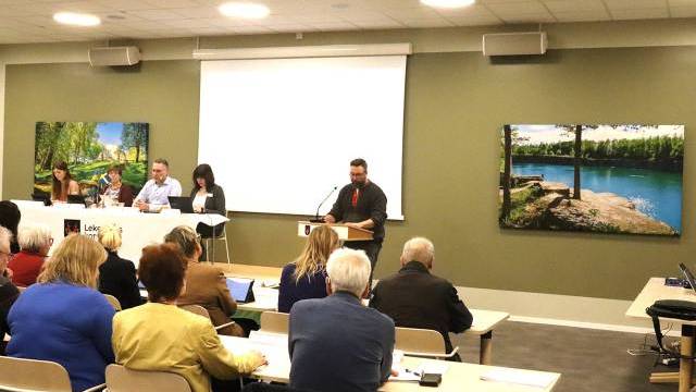 En man står på ett podium och talar till en publik, medan tre paneldeltagare sitter vid ett bord på scenen i ett konferensrum med landskapsmålningar på väggarna.