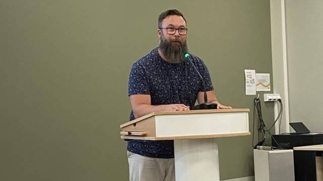 En man med skägg står på ett podium och talar i en mikrofon i ett rum med en grön vägg och elektronisk utrustning i bakgrunden.