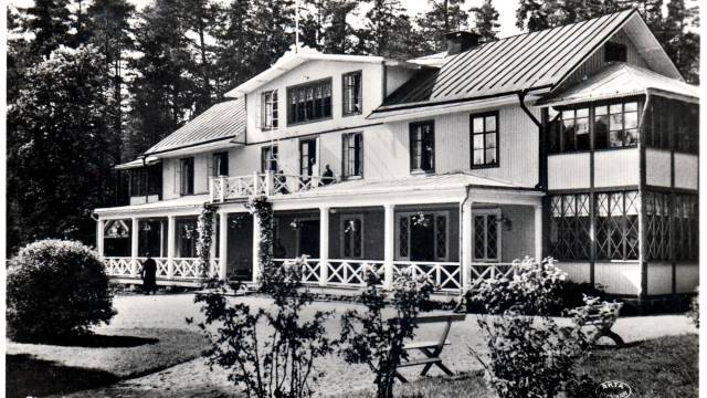 Svartvitt foto av ett stort, tvåvåningshus i trä med en veranda, omgivet av träd och buskar.