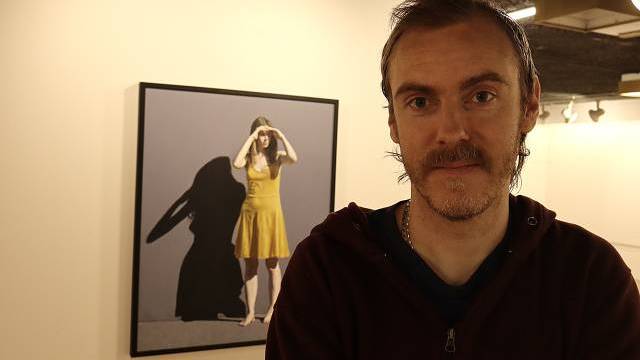 En man står inomhus med armarna i kors framför ett inramat fotografi av en kvinna i en gul klänning på väggen bakom honom.