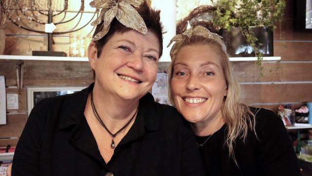 Två kvinnor med svarta toppar och pannband i bladguld ler mot varandra i ett varmt upplyst rum med växter och dekorationer i bakgrunden.